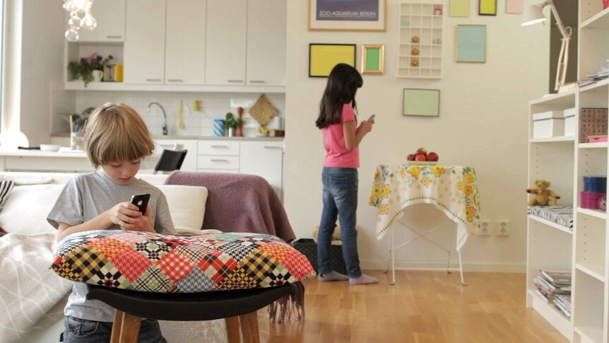 Two children stand inside their house using their mobile phones