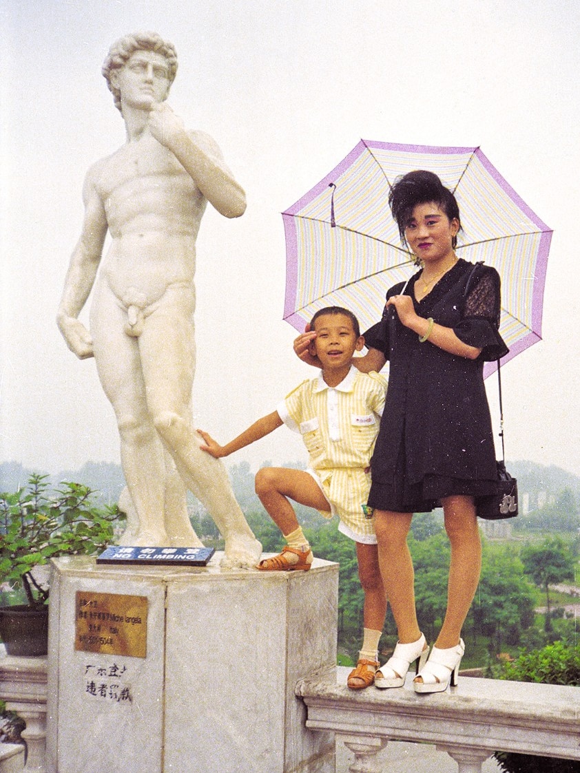 You view a woman and a young boy standing atop an Italianate balustrade next to a fake statue of Michelangelo's David.