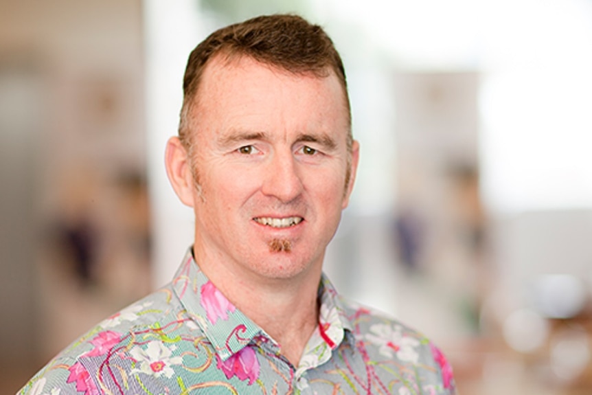 A man in a pink, white and green shirt with flowers on it with a blurred background.