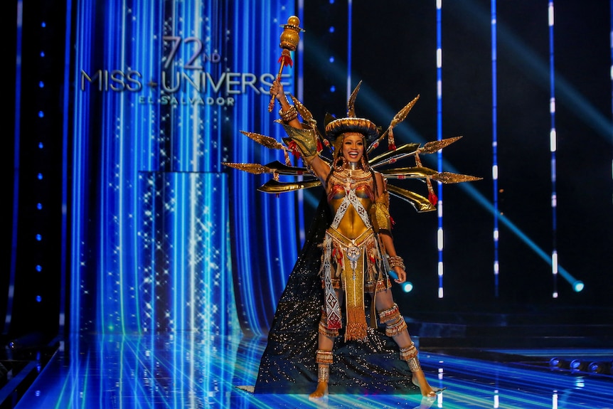 Mitchel Ihezue wearing a gold ornate bodysuit with a dark shimmery cape, gold headpiece and holding a staff