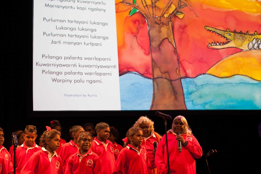 Students sing onstage in front of a screen with a drawing of a crocodile and tree and a song in Walmajarri