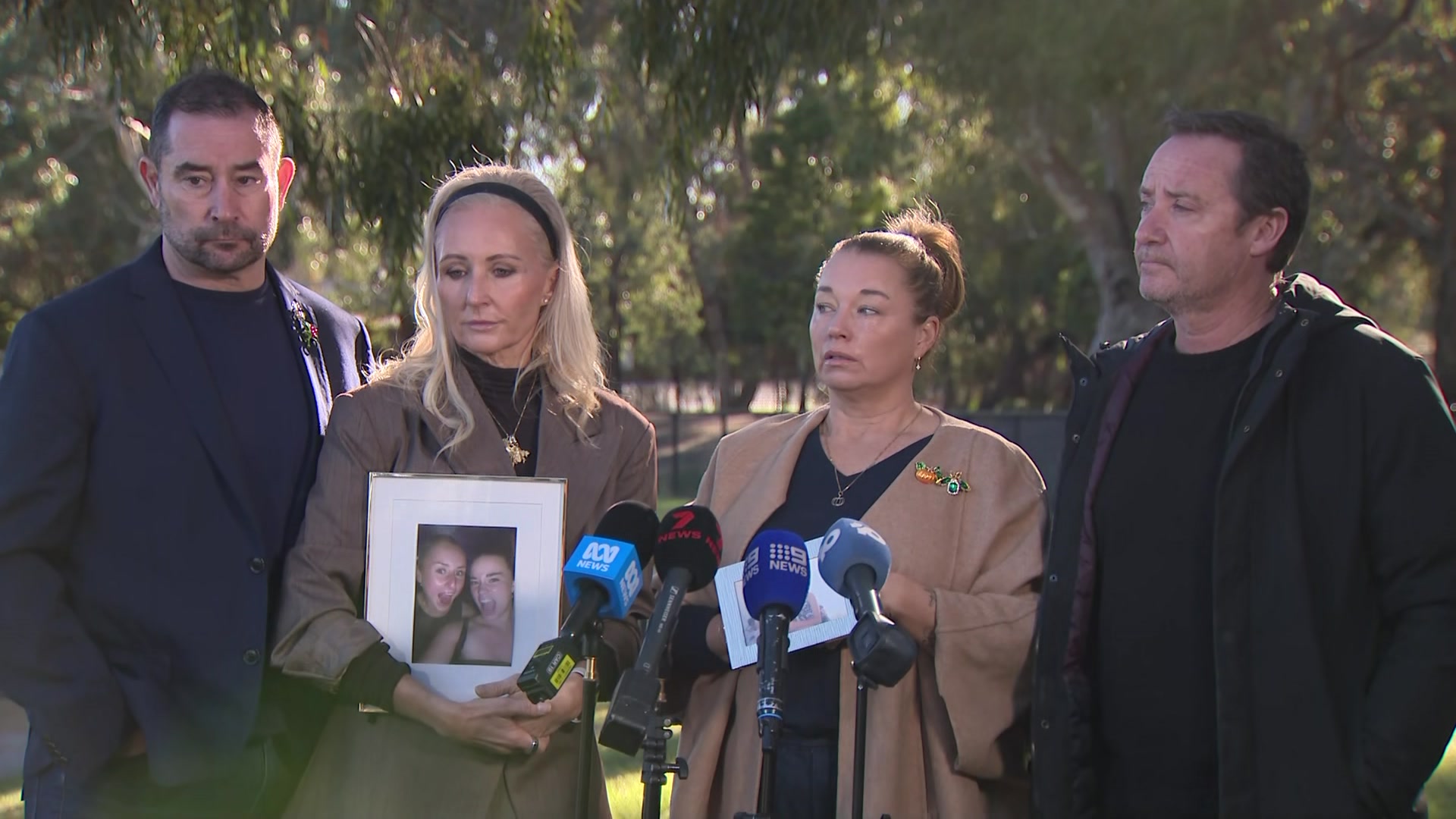 Two  men in dark jackets and shirts booked two women in beige coats and black tops who stand behind microphones.