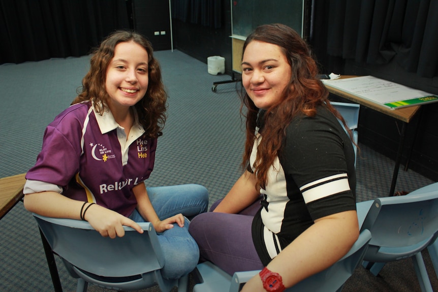 Two high school students sitting on chairs in their drama room, smiling.