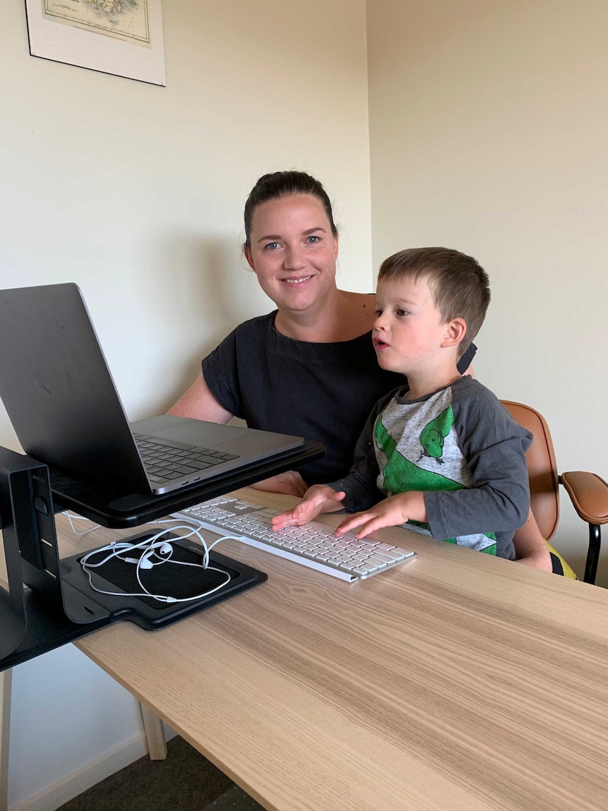 Tory sits at her desk smiling while Isaac looks with interest at her computer.