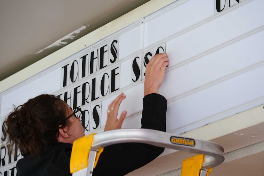 A woman on a ladder removes letters from a noticeboard at a cinema.