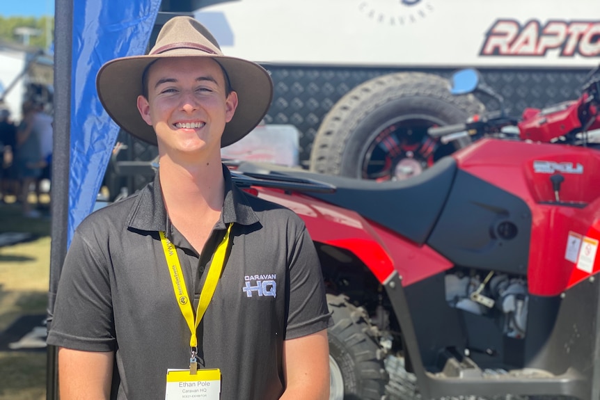 Young man with black polo shirt on and wide brim hat standing next to a caravan 