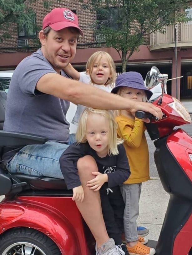 Three small children, triplets, stand on the base of a red electric scooter where their dad sits on the seat