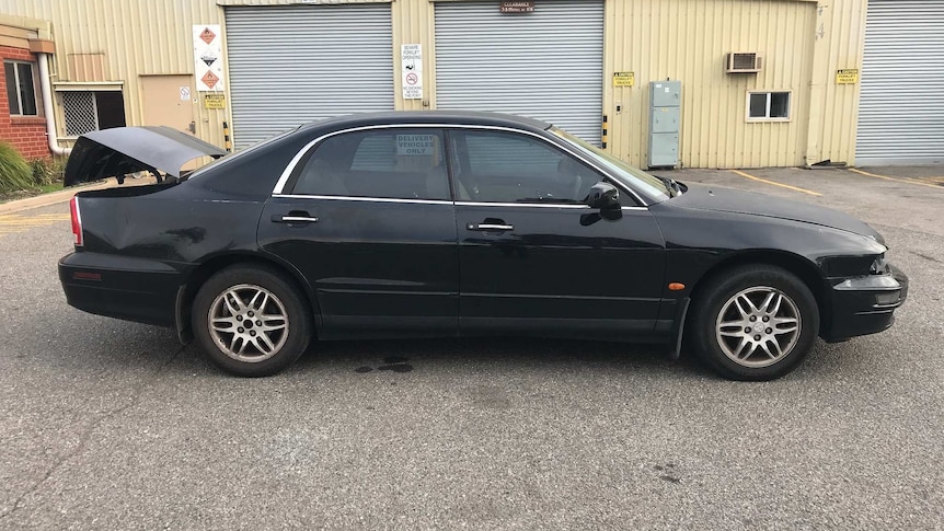 A black sedan with its boot open in front of a large shed