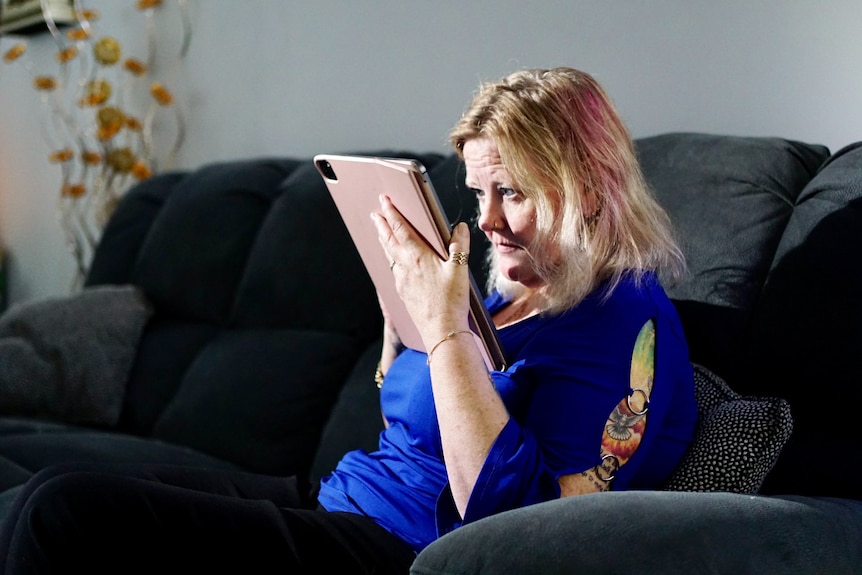 A woman sits on a couch and reads from a digital tablet held close to her face.