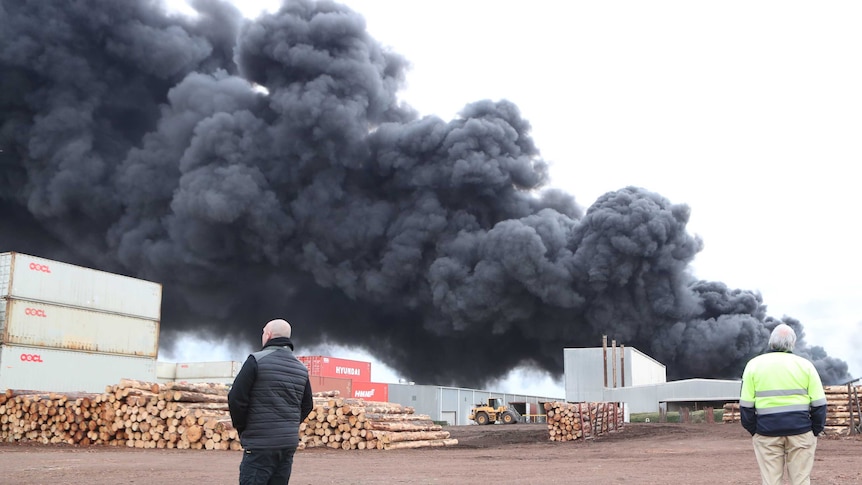 Men at a timber yard walking in the foreground of a huge plume of smoke.