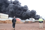 Men at a timber yard walking in the foreground of a huge plume of smoke.