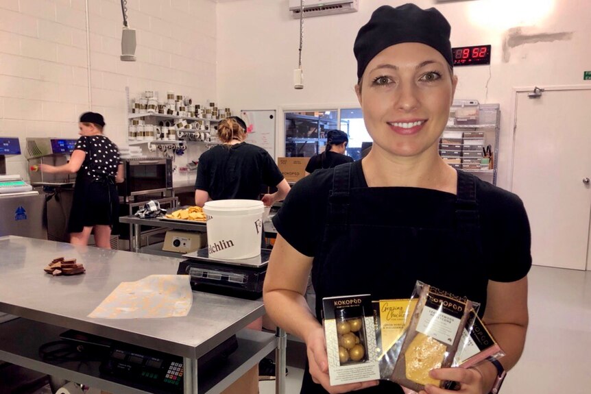 A lady wearing a black hair cap smiles at the camera holding a range of chocolates with workers behind her.