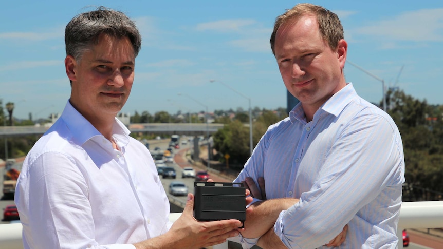 Martyn Gilbert and Adam Boyt stand on a bridge holding their Sentinel emergency device, with a Perth freeway in the background