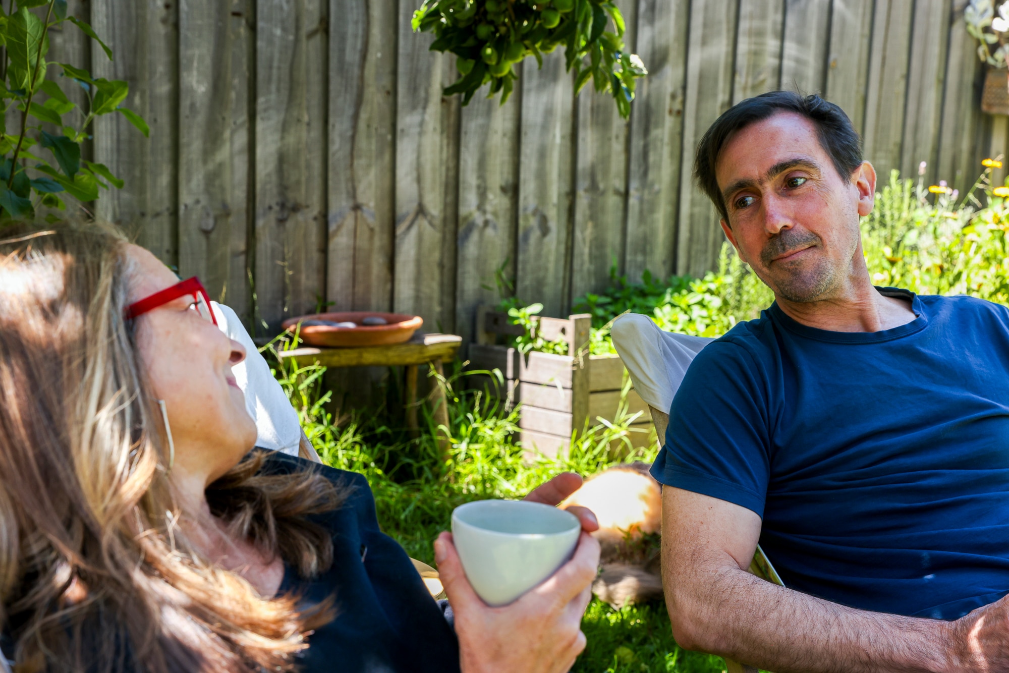 A man and a woman sit outside. They are leaning back in their chairs looking at each other lovingly. She holds a cup,