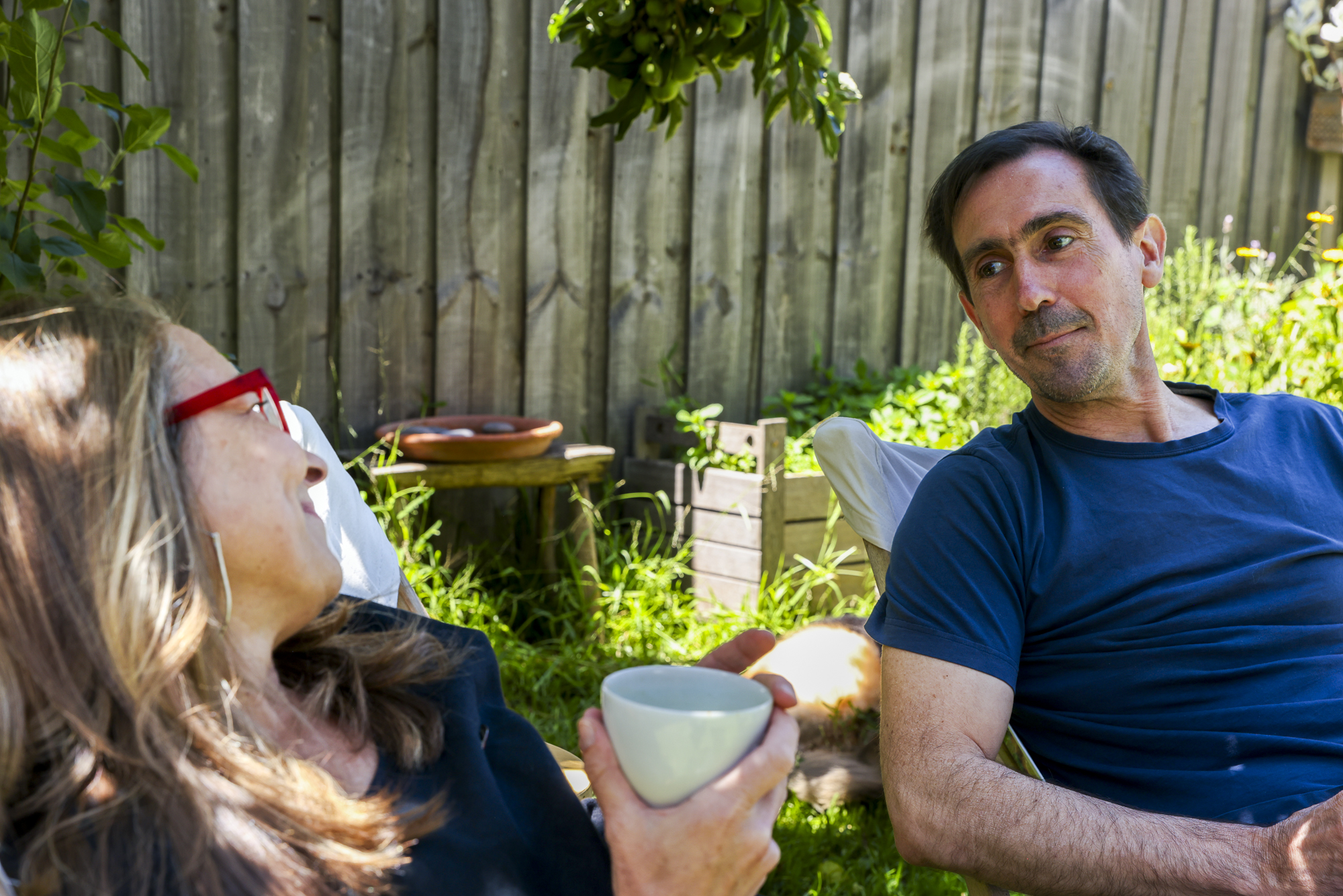 A man and a woman sit outside. They are leaning back in their chairs looking at each other lovingly. She holds a cup,