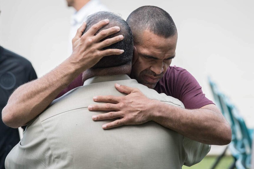 Anthony Mundine, wearing a brown tshirt, embraces a man wearing a beige jacket.
