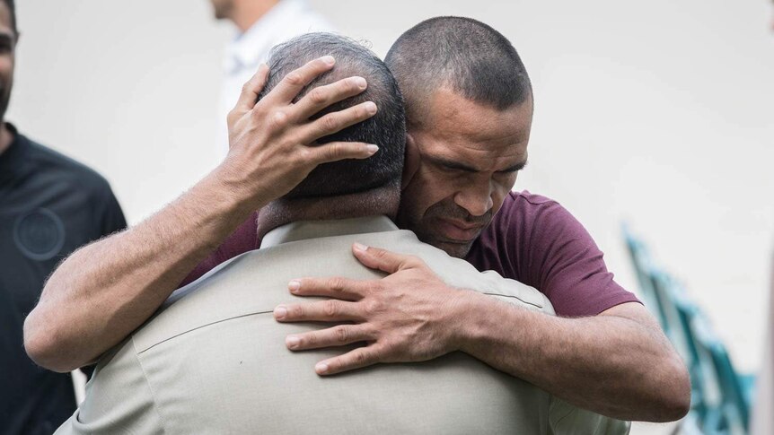 Anthony Mundine, wearing a brown tshirt, embraces a man wearing a beige jacket.