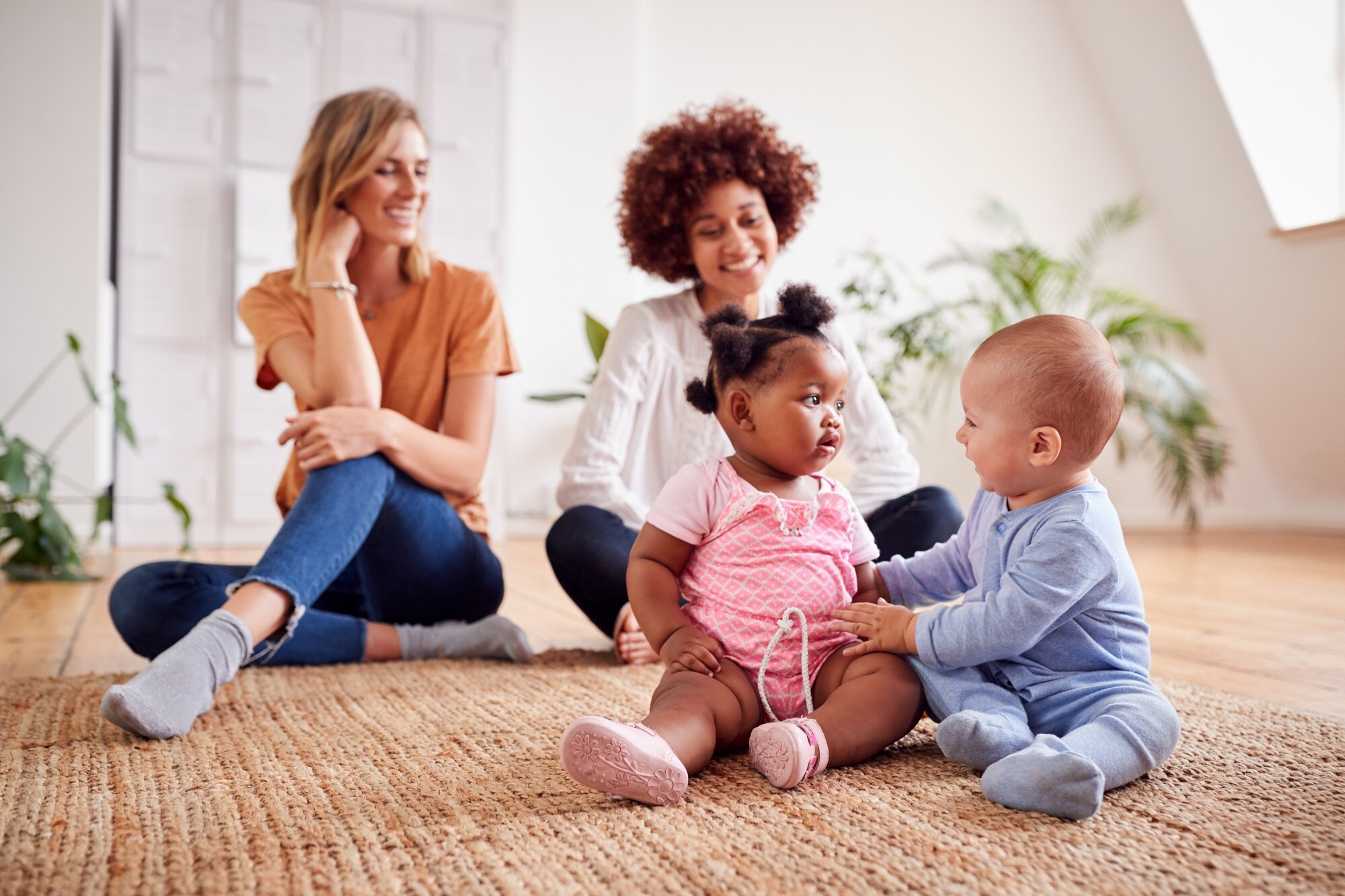 Two toddlers playing with each other, their mums watching and smiling in the background