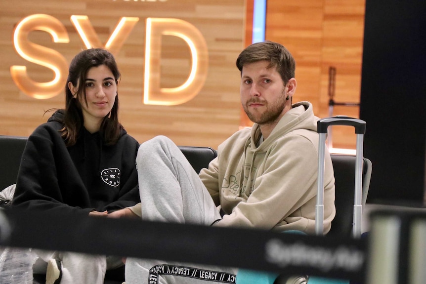 A young woman and man sitting in front of the Sydney Airport departures sign