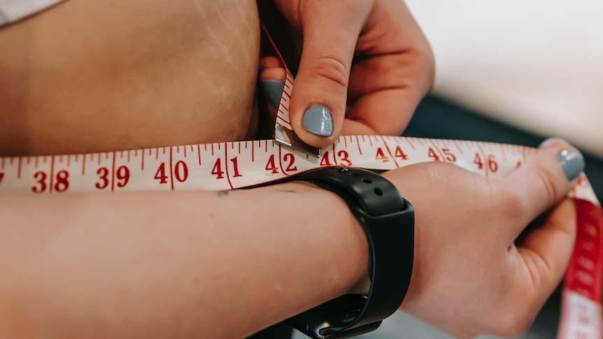 A woman wearing a black wrist watch and grey nail polish measures her waist with a red and white measuring tape
