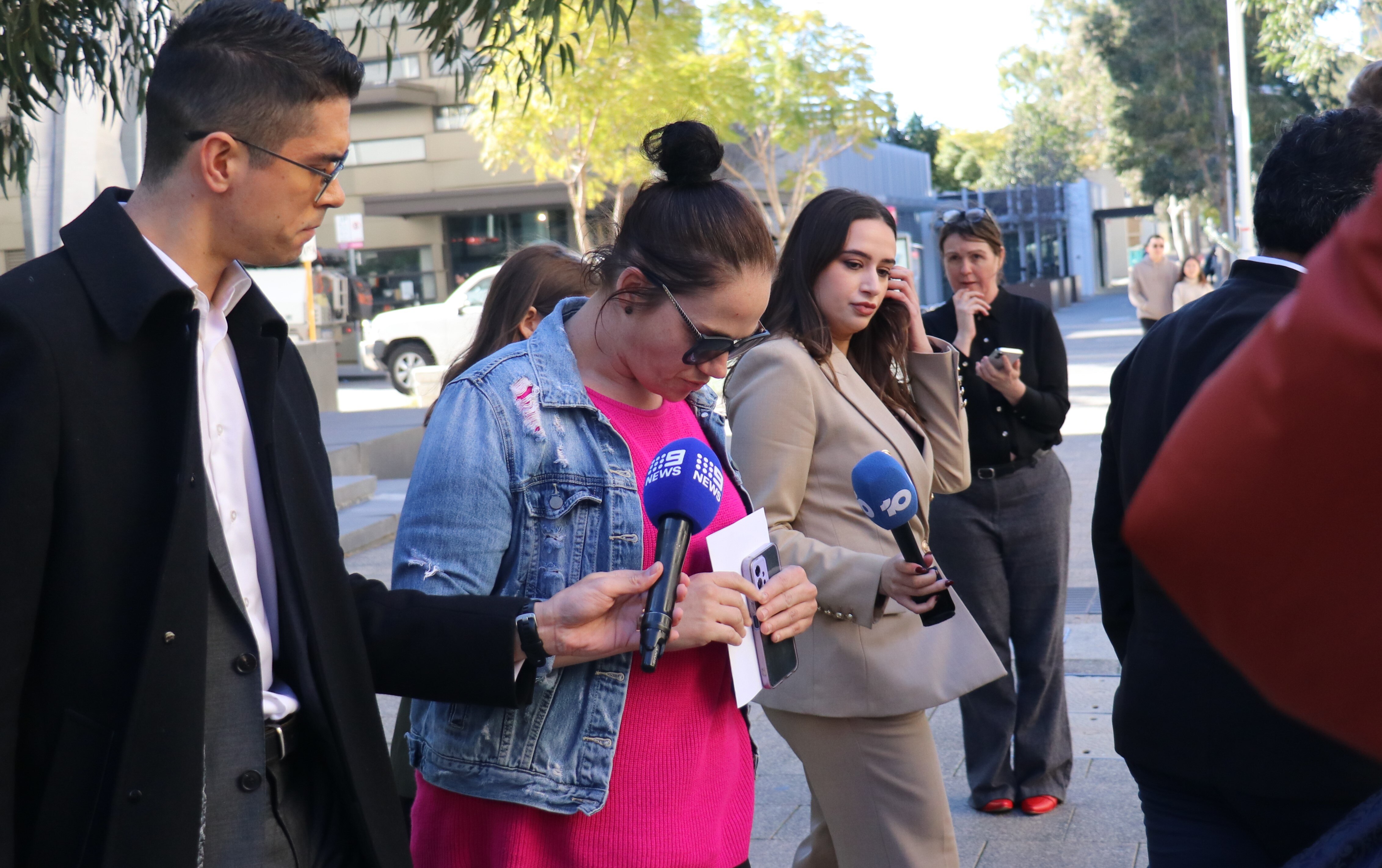 A woman with pink top and denim jacket with head down as reporters point microphones towards her  