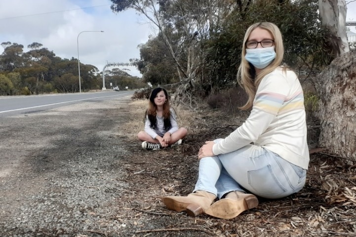 A woman wearing a face mask sits on the side of a highway with a girl behind her