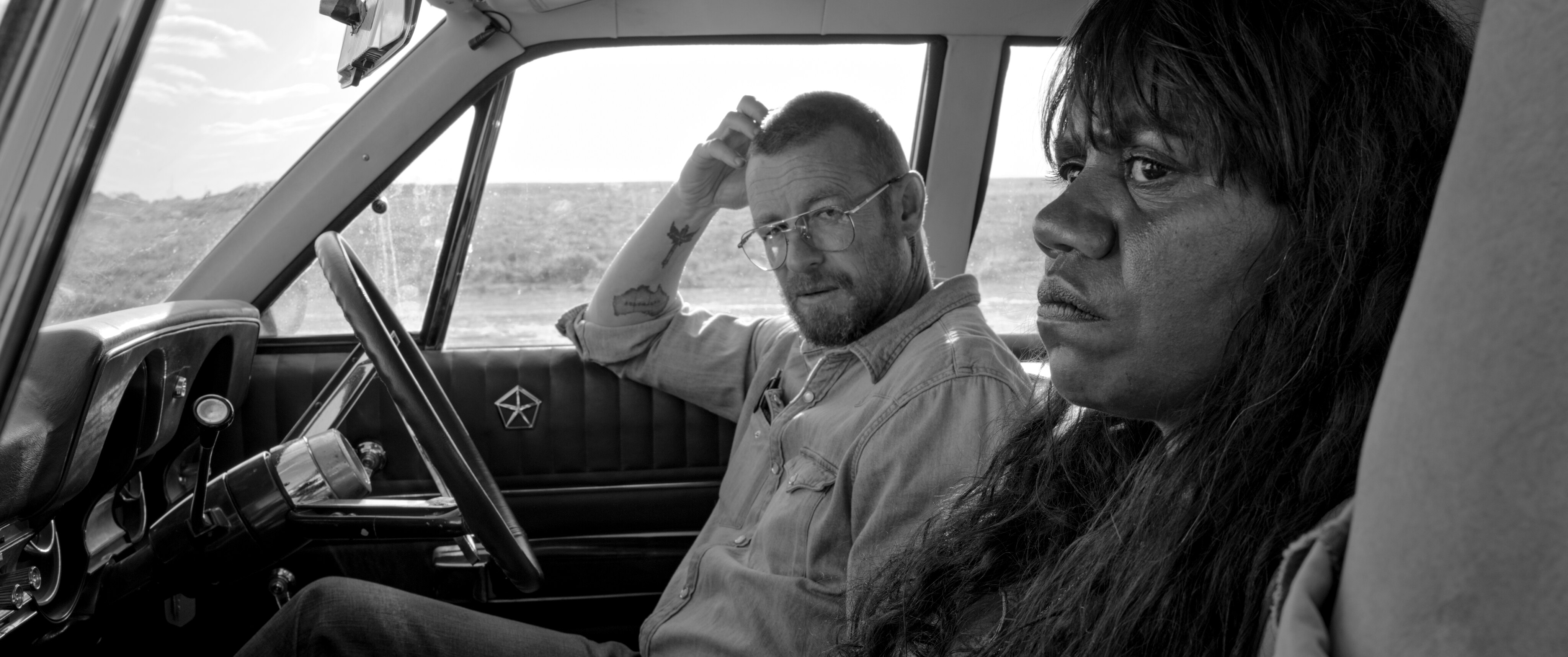 A black and white photo of a white man with glasses and a beard in a car, looking at his female Indigenous passenger.