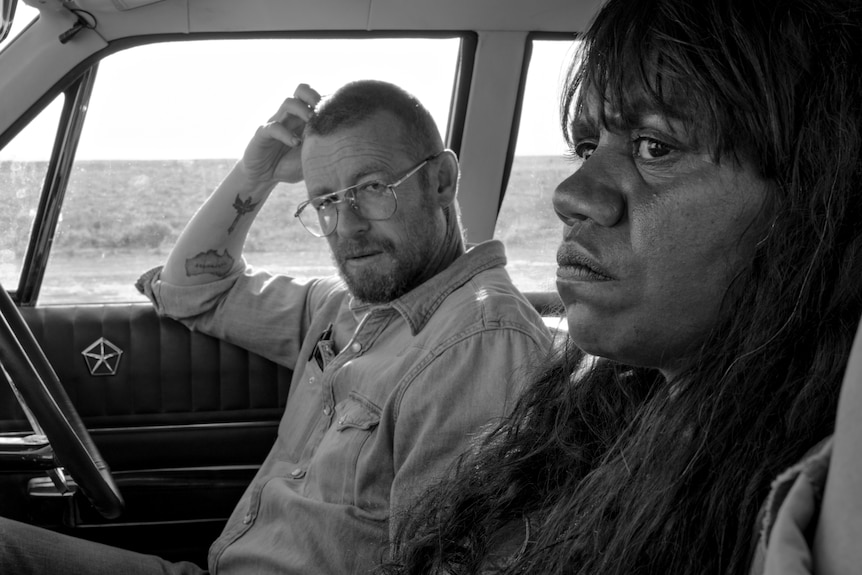 A black and white photo of a white man with glasses and a beard in a car, looking at his female Indigenous passenger.