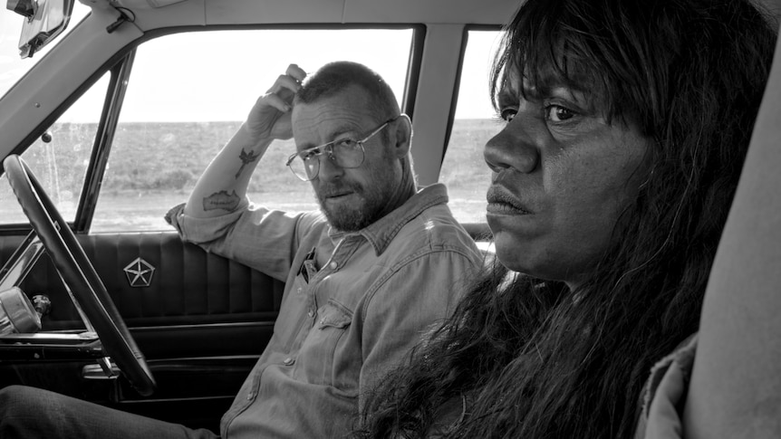A black and white photo of a white man with glasses and a beard in a car, looking at his female Indigenous passenger.