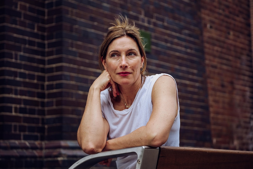 Woman in a white T shirt looks to her left leaning on a red brick wall