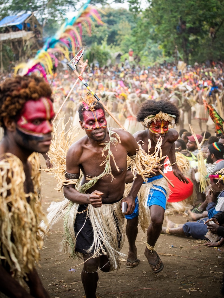 Two men running wearing facepaint and grass skirts over shorts with many people behind them.