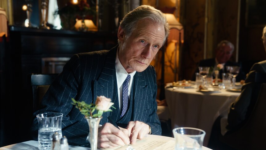 A 70-something man in 50s garb leans forward over a table in a restaurant, with a stony but sad expression