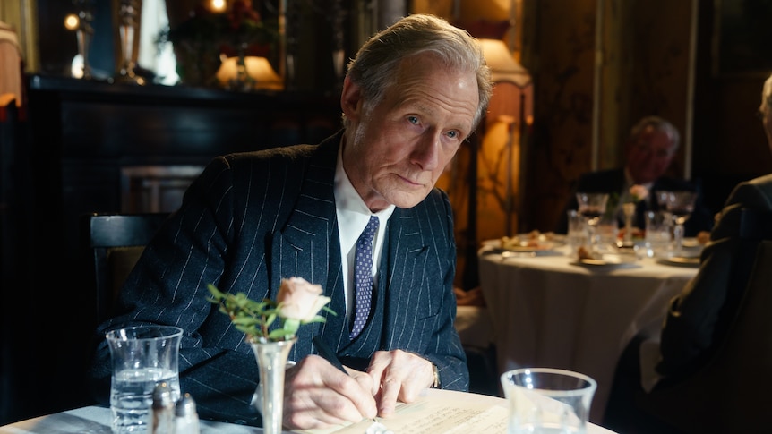 A 70-something man in 50s garb leans forward over a table in a restaurant, with a stony but sad expression