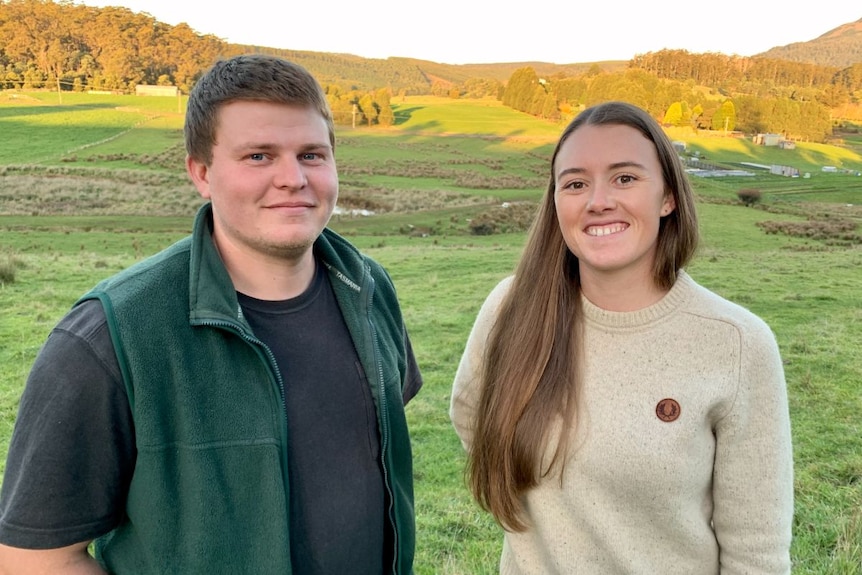 A man in a green vest and t shirt and a woman in a beige jumper stand in a green paddock.