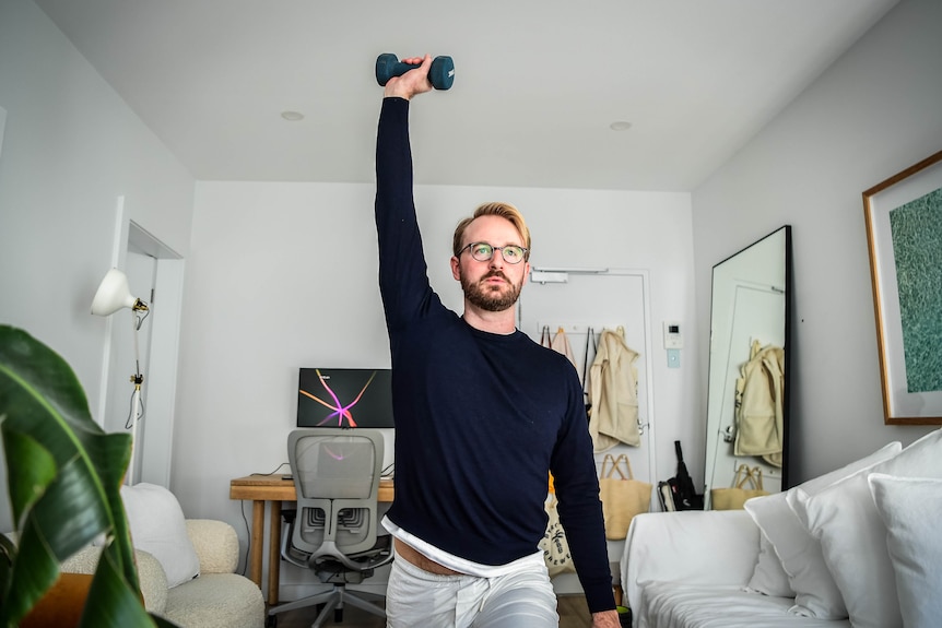 Giles holds a dumbbell above his head with one hand. His home-office set-up, including desk and computer, is in the background.