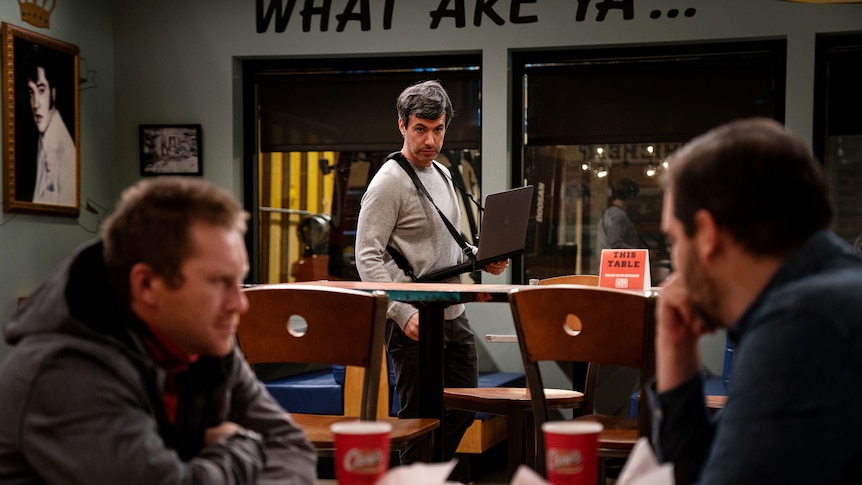 Comedian Nathan Fielder wearing a laptop in a harness while looking at two people in the foreground