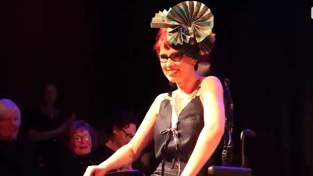 Woman sits in a wheelchair under bright lights wearing a hair piece, glasses and a black jumpsuit.