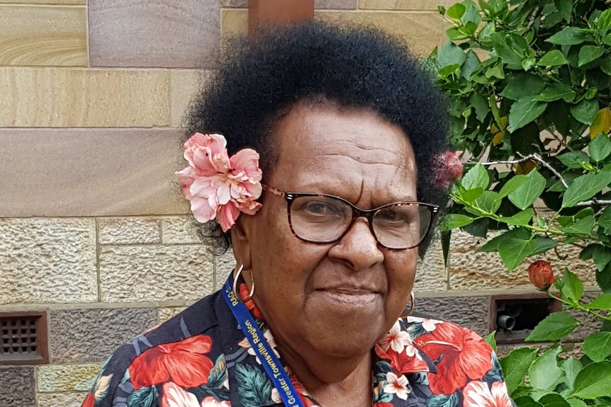 A woman, wearing glasses, a flower in her hair and a floral shirt, smiles at the camera.