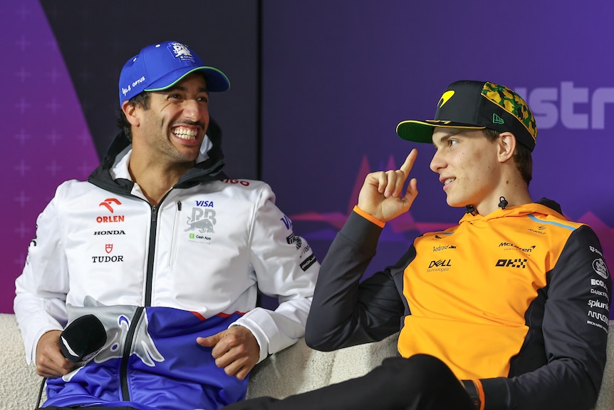 Daniel Ricciardo and Oscar Piastri sitting on a couch, during a media conference, wearing team merchandise