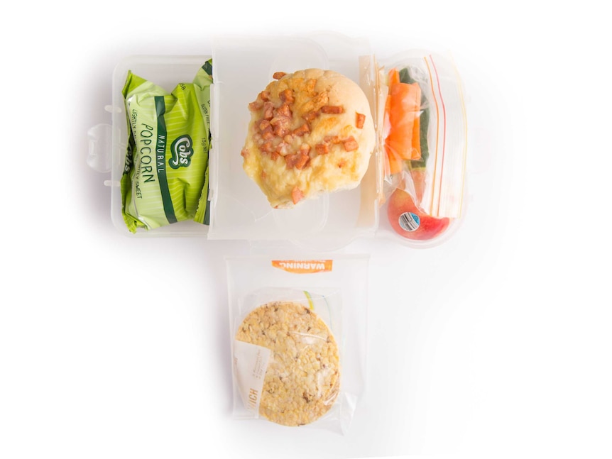 A cheese and bacon roll, popcorn, rice crackers and chopped vegetables and fruit in a lunch box.