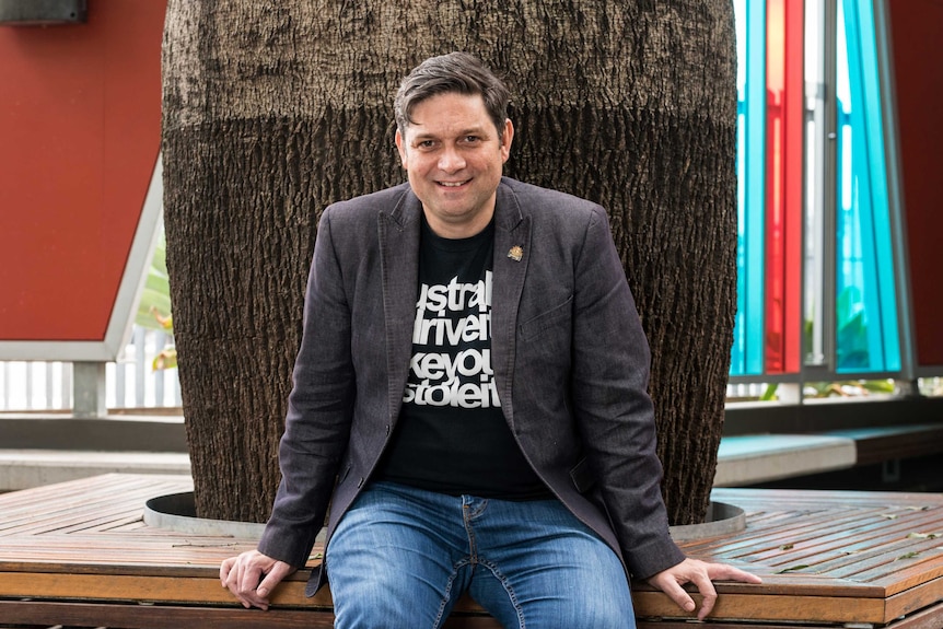 Colour photograph of Sydney Festival artistic director Wesley Enoch sitting in on a bench outside.