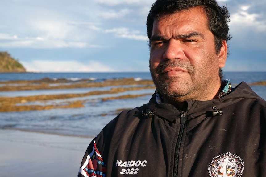 Close-up of man standing on a beach, with a rocky shoreline and the ocean behind him