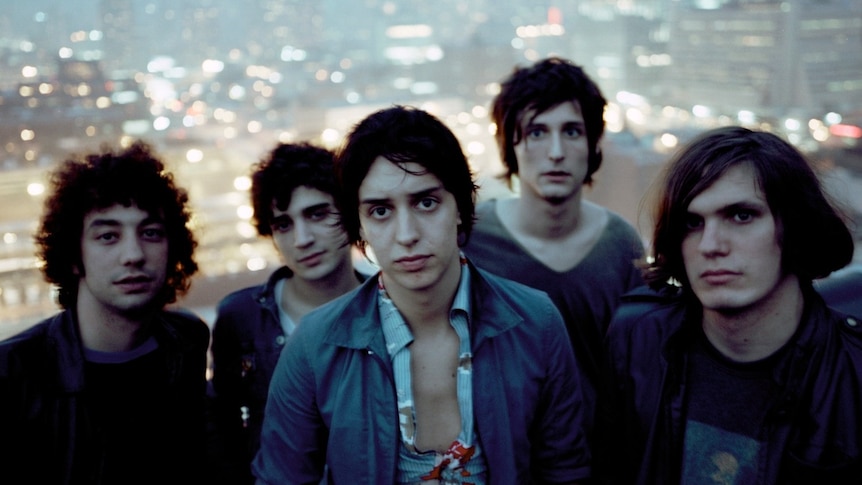 A slightly blurry portrait of the five members of The Strokes atop a building with the New York City skyline in the background