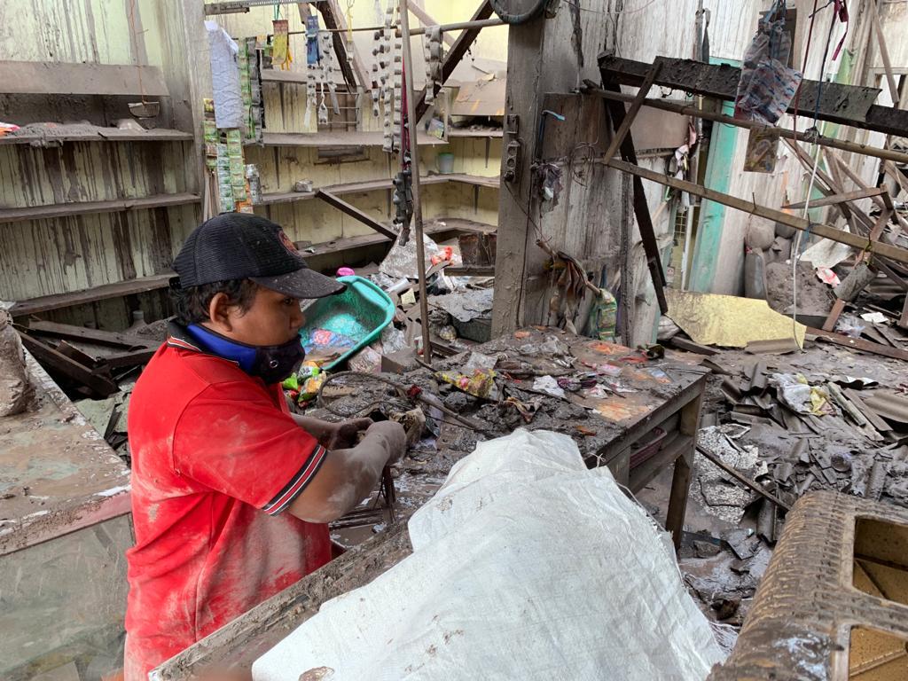 A man in a red shirt and a black cap in the middle of the debris of his house.