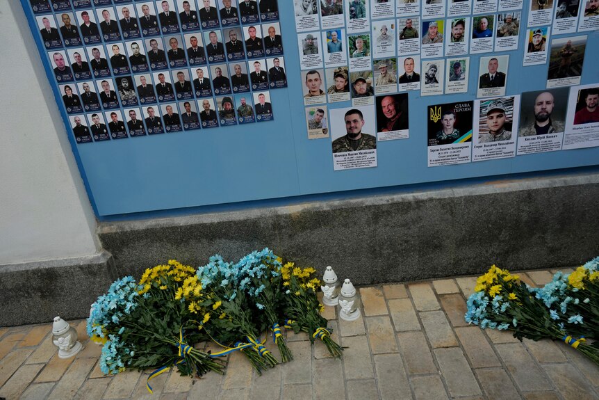 Blue and yellow flowers rest below a wall with pictures of Ukrainian soldiers killed in the war.