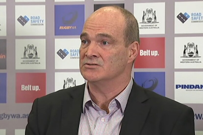 A mid shot of Mark Sinderberry wearing a suit and talking in front of a pop-up display showing Western Force sponsors.
