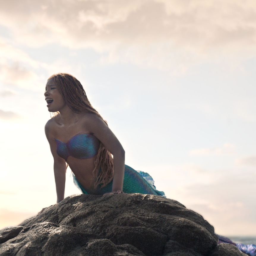 A mermaid perches upon a rock, she has dark skin and hair and is singing