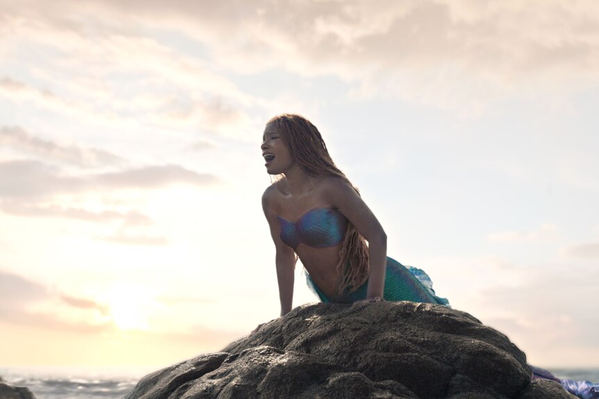 A young black woman with red-toned dreadlocked hair lifts her body up onto a submerged rock in the ocean, singing.