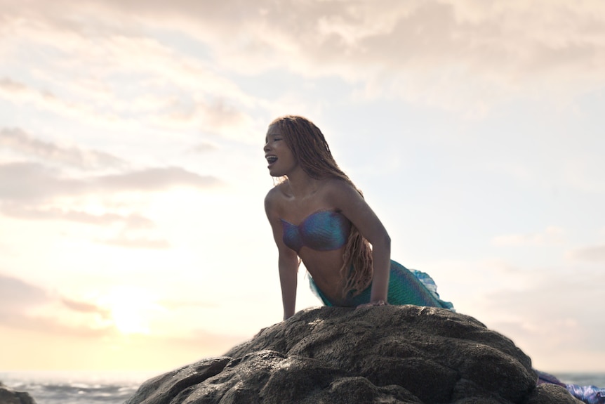 A young black woman with red-toned dreadlocked hair lifts her body up onto a submerged rock in the ocean, singing.