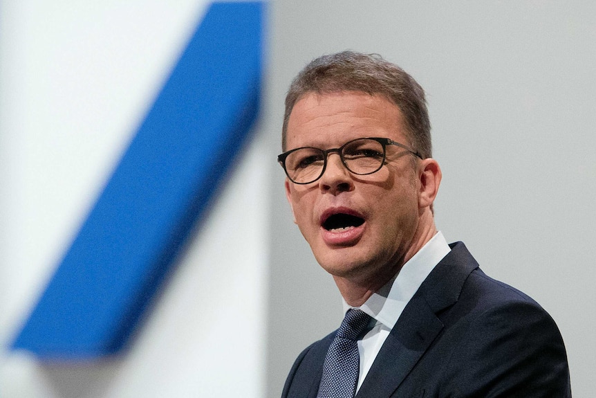 A man in a navy suit and tie is pictured close up with his mouth open in front of a blue slash — the Deutsche Bank logo.